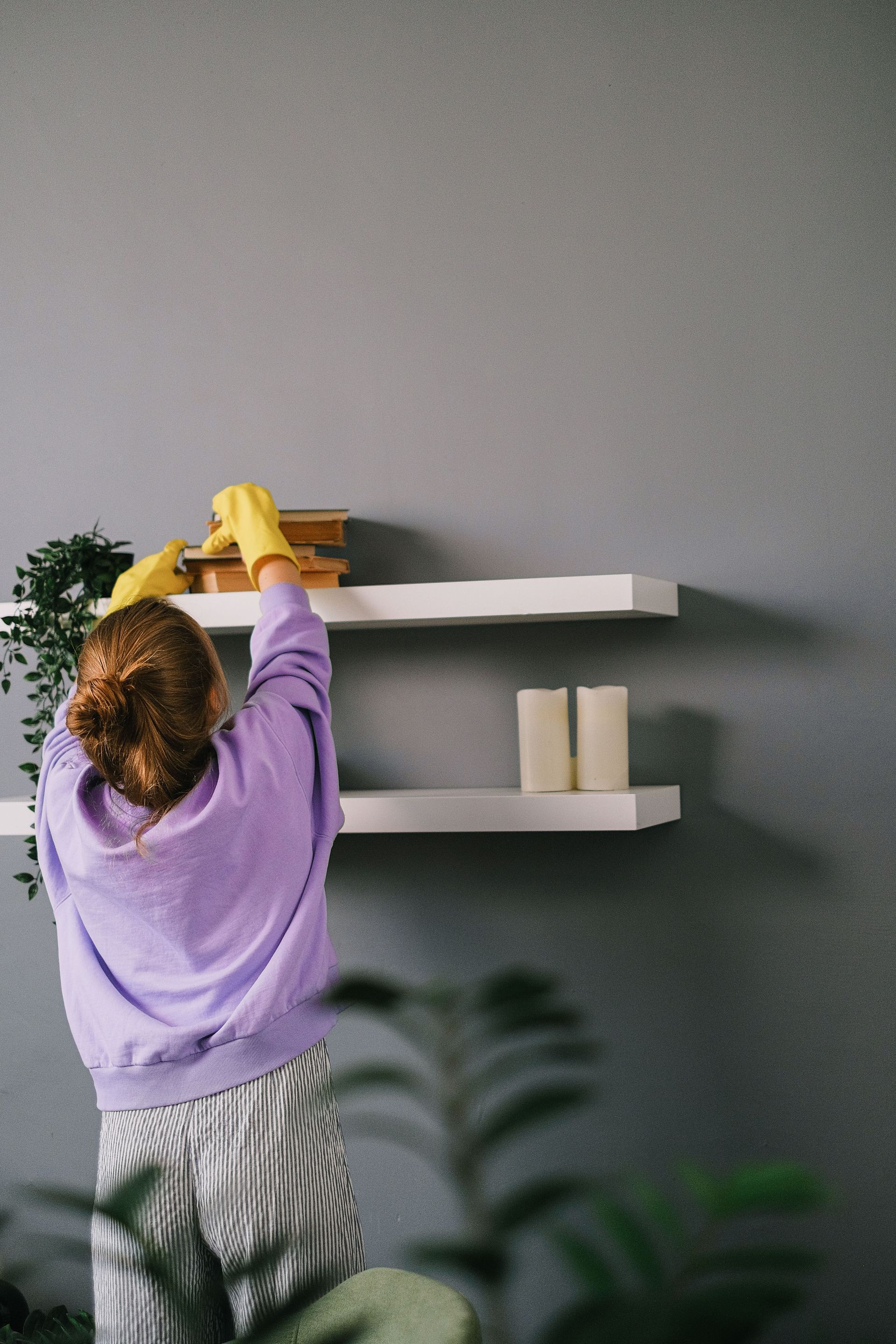 Back view of unrecognizable female in casual clothes and latex gloves standing and taking books while cleaning shelves in room with gray wall